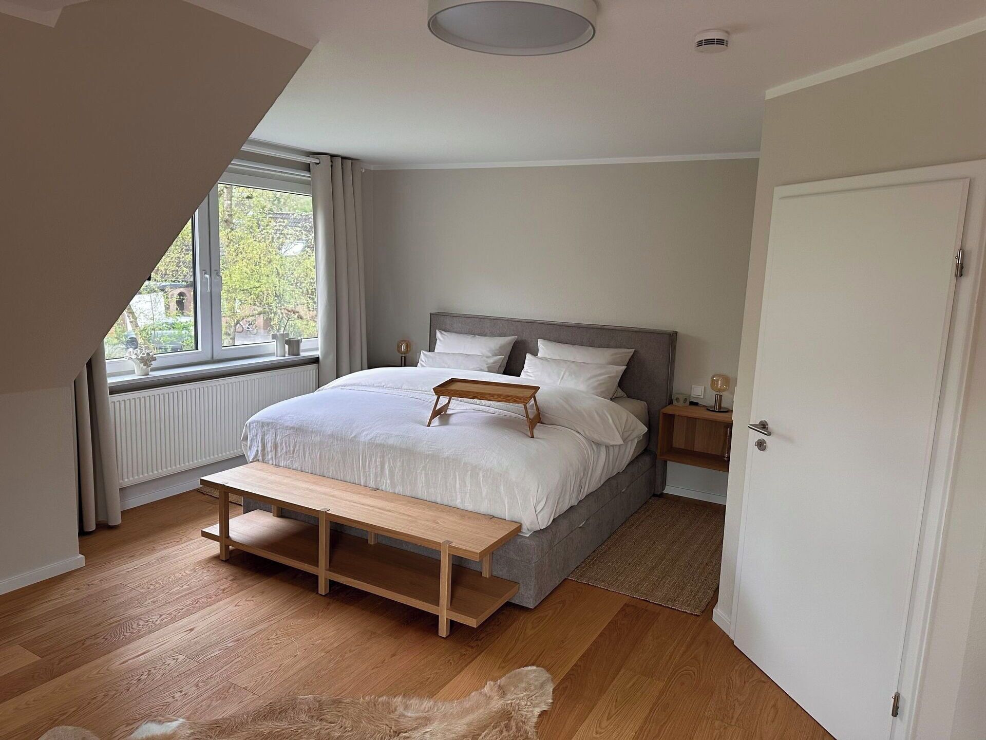 Une chambre à coucher comprenant un lit, des tables de chevet, un banc, un radiateur, une fenêtre avec des rideaux et une porte.