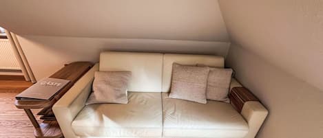 A corner room with a white leather sofa, two pillows, a wooden side table, and a book titled 'GÄSIE'.