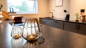 A kitchen counter with two glass candle holders, a window with blinds, and a sink. - North Nest Signature Apartment (Tromsø)