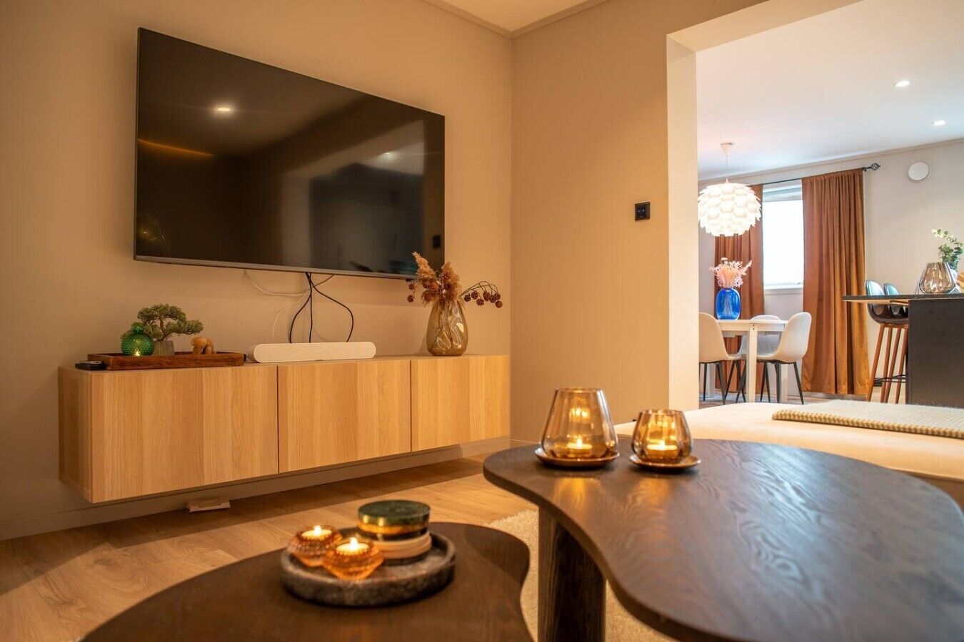 A modern living room with a flat-screen TV mounted on the wall, a wooden console table, and a dining area with a table and chairs.