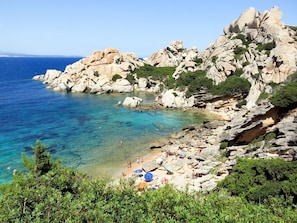 Eine felsige Küstenlinie mit klarem, blauem Wasser, einem Sandstrand und üppigem Grün.