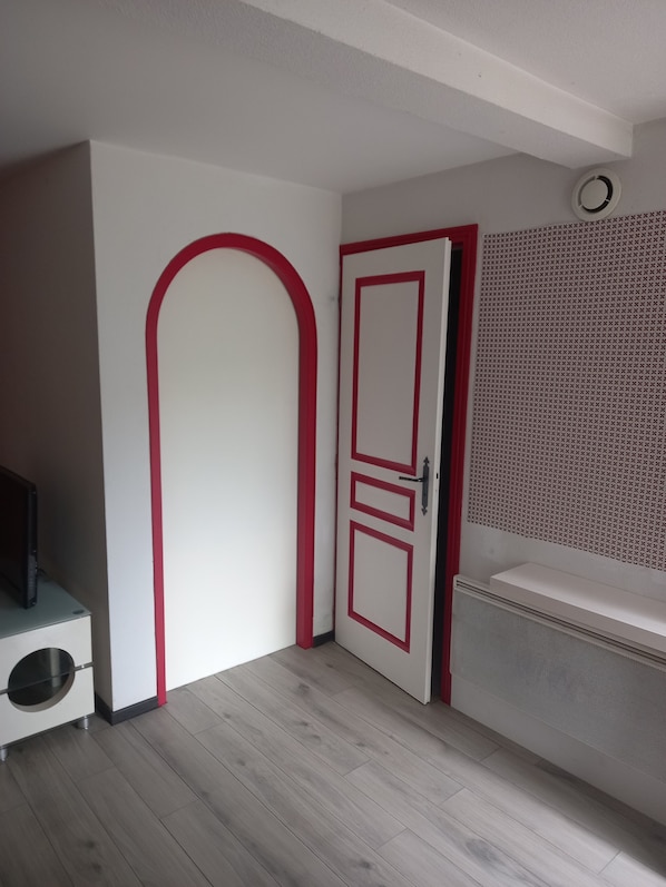 Une pièce avec une porte blanche bordée de rouge, un meuble blanc sur lequel repose un objet noir, et un évier blanc surmonté d’un panneau perforé.