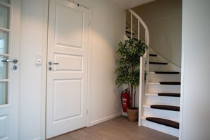Eine Treppe mit weißer Handläufung und Holzboden, eine weiße Tür mit Schlüsselloch und ein Topfpflanzen neben einem Feuerlöscher.