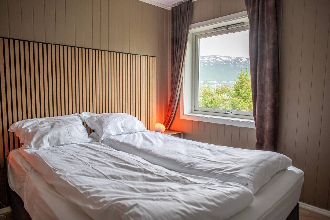 A neatly made bed in a room with a window overlooking a mountainous landscape.