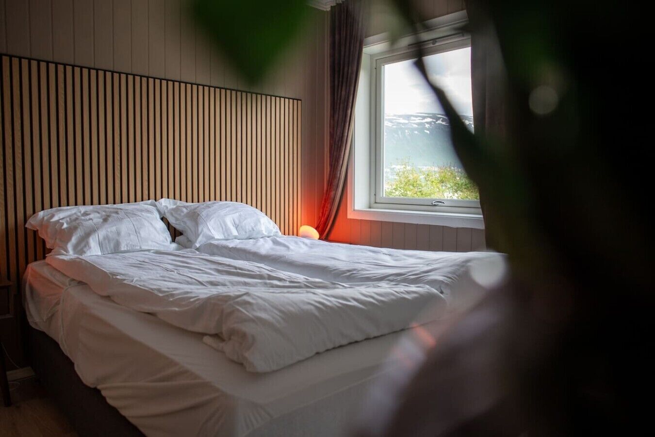 A bed in a room with a slatted headboard, a window with curtains, and a view of greenery outside.