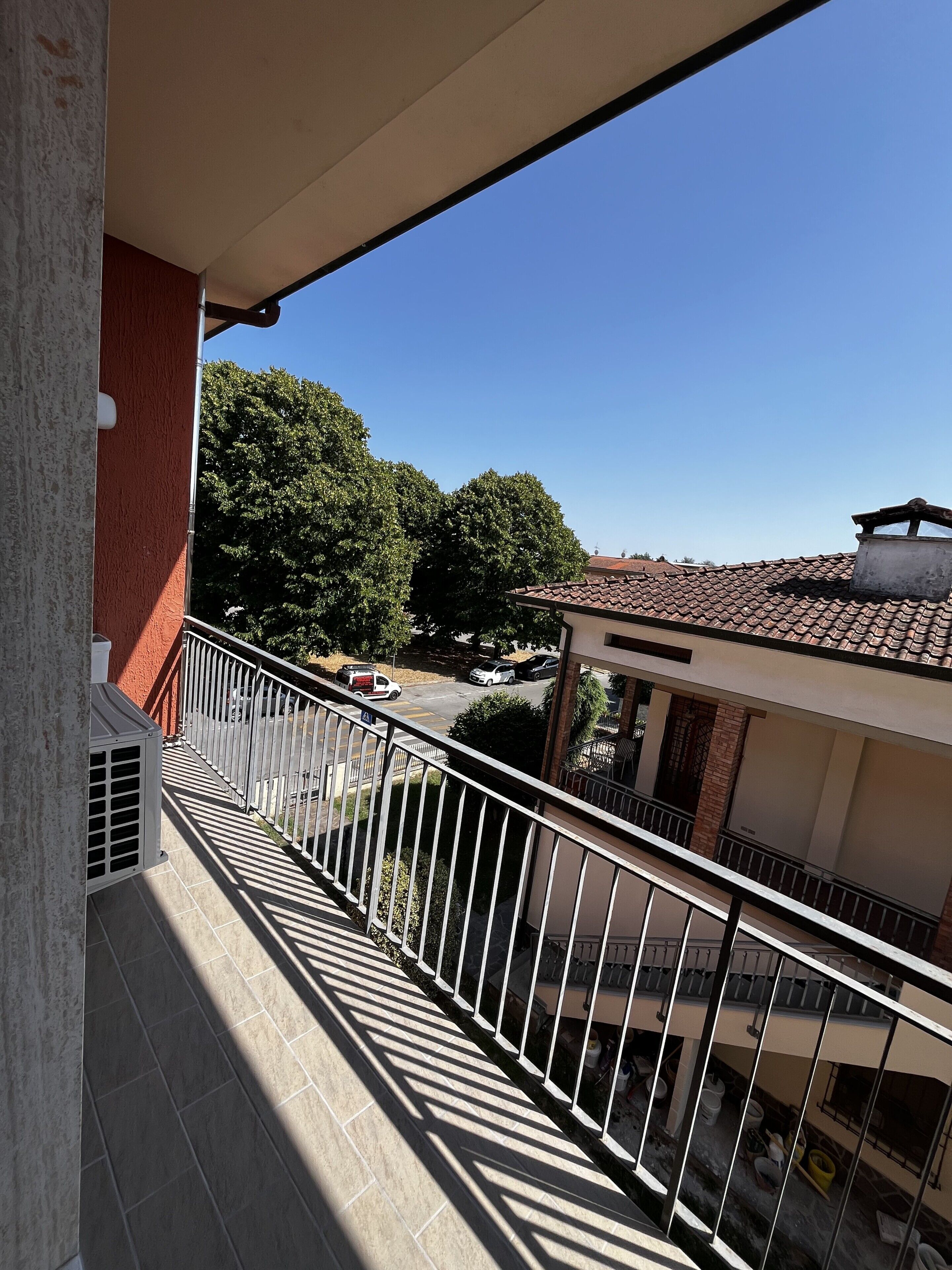 A balcony with a view of a street, a building with a tiled roof, and a tree.