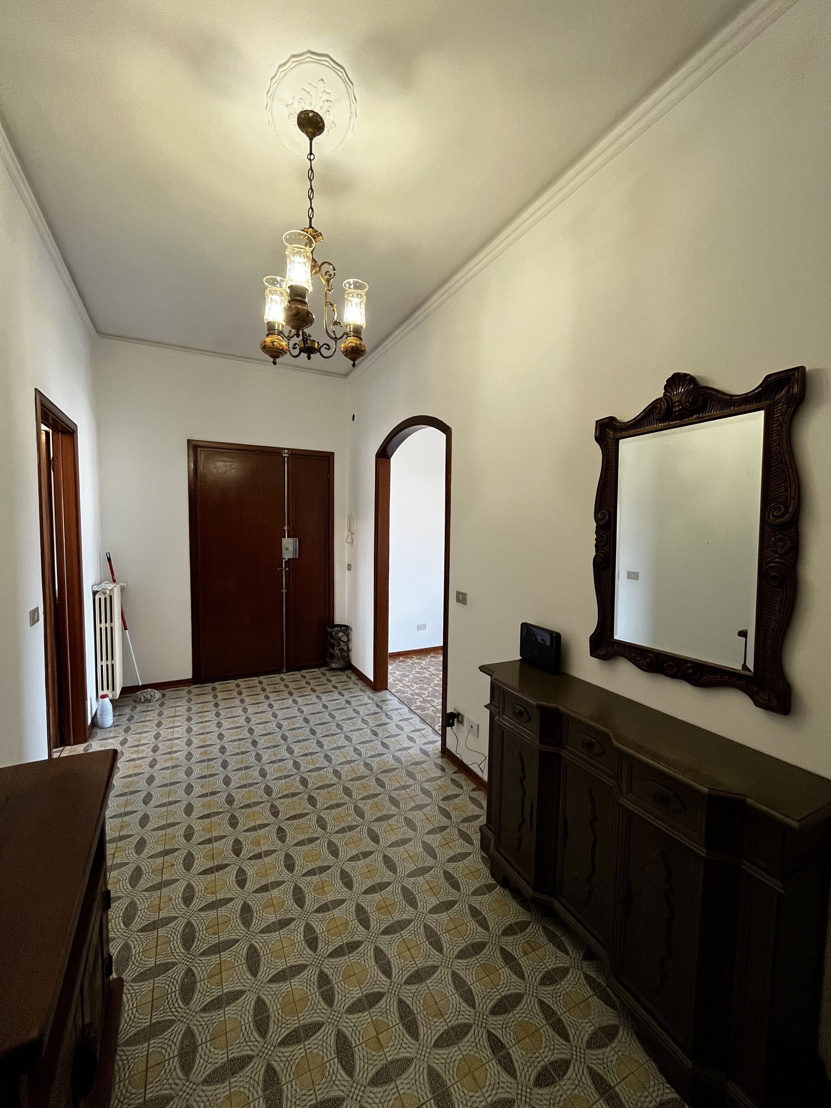 A hallway with patterned floor tiles, a wooden door, a mirror on the wall, and a chandelier.