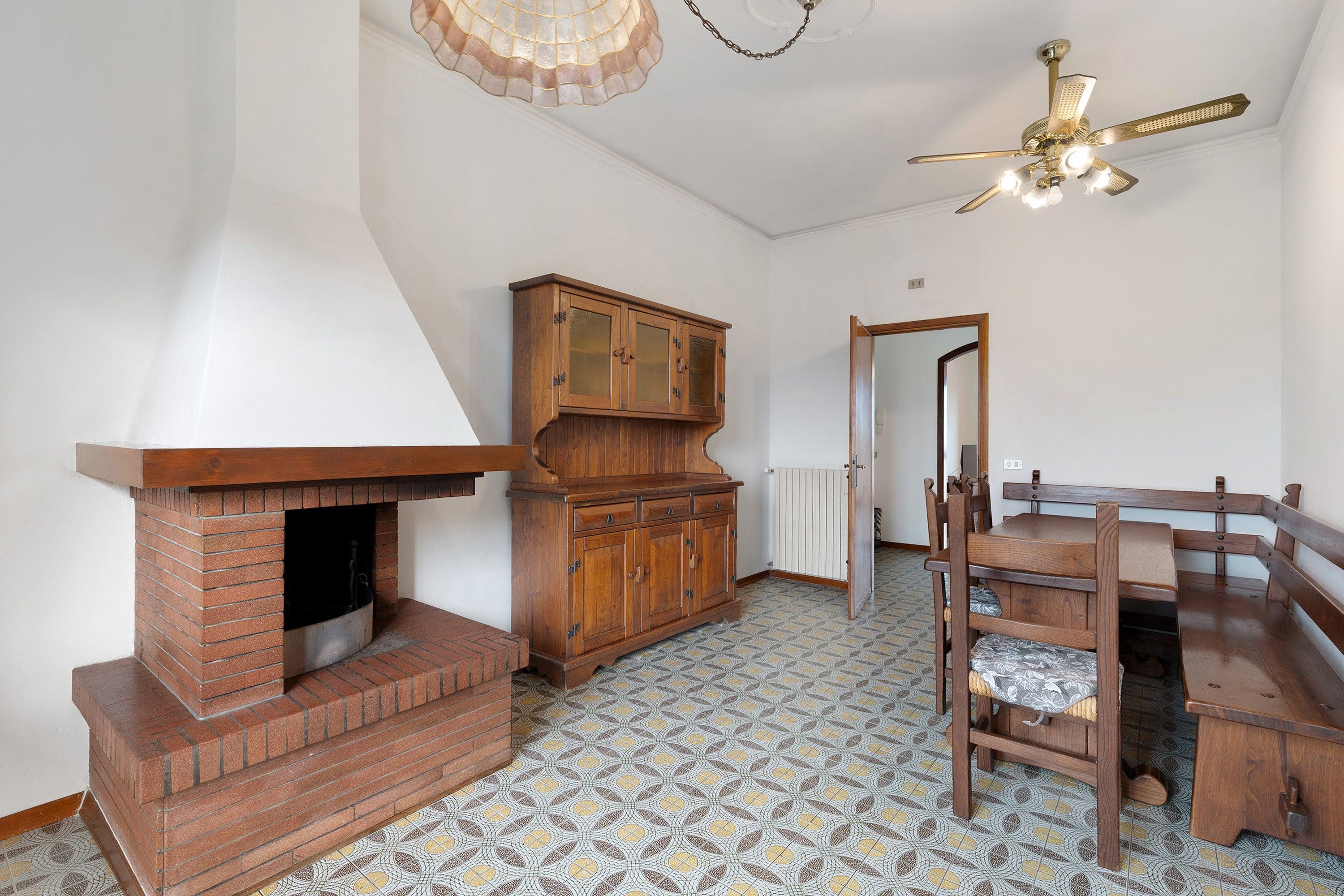 Ein Zimmer mit Kamin aus Backsteinen, Holzmöbeln, gemusterten Bodenfliesen und einem Deckenventilator.