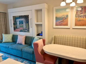 A living room with a blue sofa, a red chair, a white table, and two framed pictures on the wall. - Orlando | Lively Group Stay | Pool + Walkable BoardWalk | 2BR Lock-Off (Lake Buena Vista)