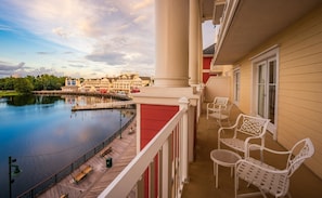 A balcony with white chairs and a view of a calm lake and hotel buildings. - Orlando | Lively Group Stay | Pool + Walkable BoardWalk | 2BR Lock-Off (Lake Buena Vista)