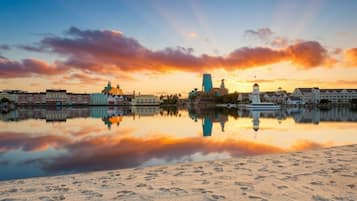 Paesaggio urbano con edifici che si riflettono in acque calme, un faro e una spiaggia sabbiosa.
