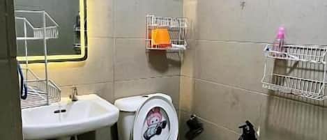 Baño pequeño con inodoro, lavamanos y espejo. Hay repisas para guardar cosas en la pared y una zona de ducha.