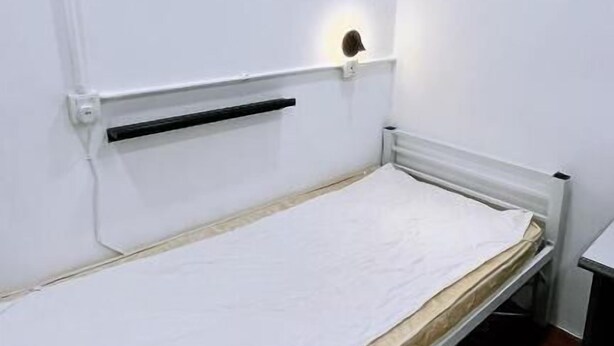 Quarto pequeno com uma cama de solteiro, colchão branco, cabeceira com luminária e radiador abaixo.