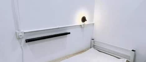 Une petite chambre avec un lit simple, un matelas blanc, une tête de lit dotée d’un luminaire et un radiateur en dessous.