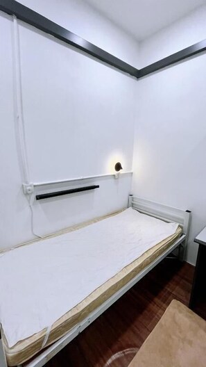 A small, single-bed room with a white mattress, a headboard with a light fixture, and a radiator below.