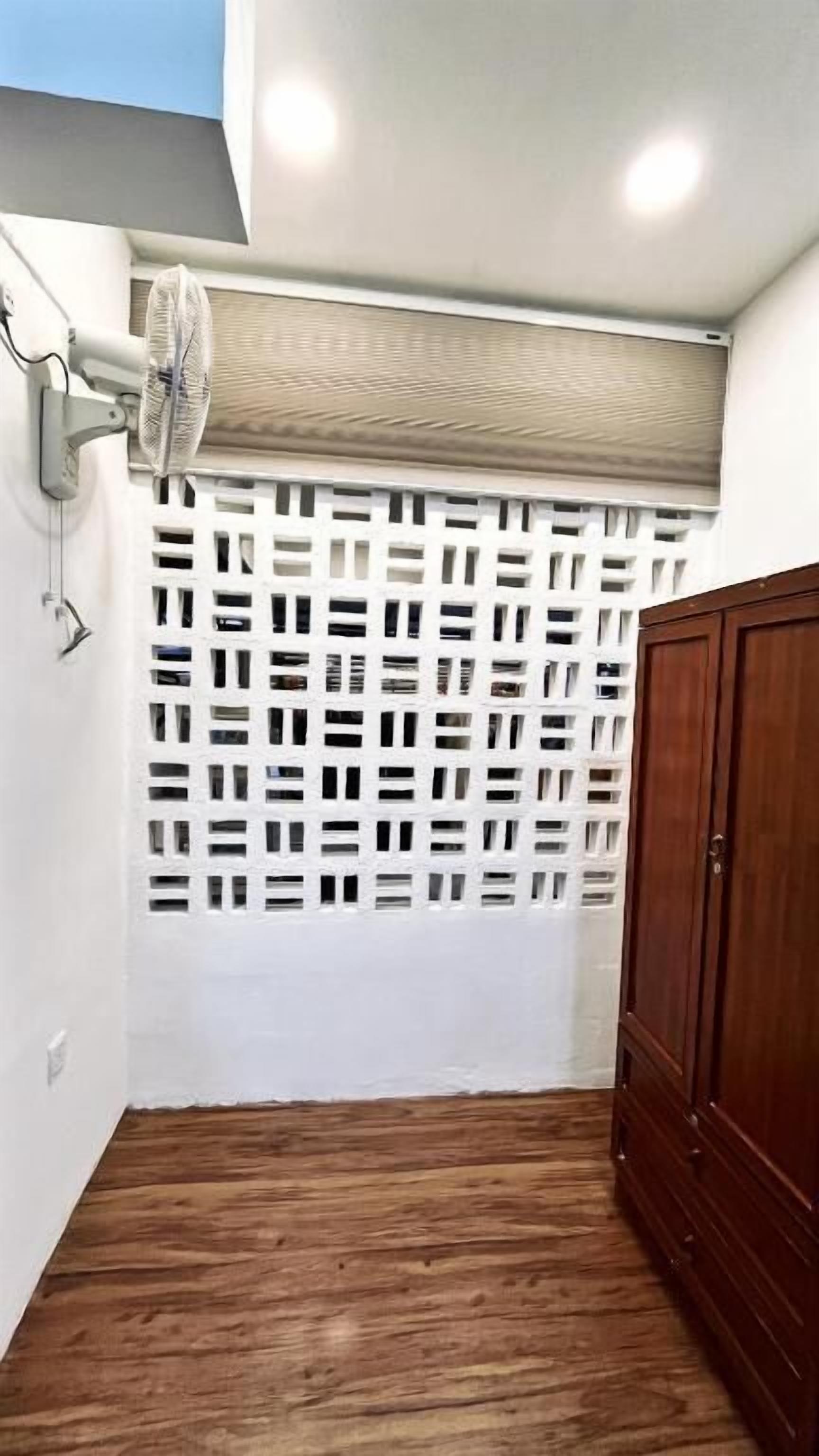 Une pièce avec un plancher en bois, un mur blanc, une porte en bois, un ventilateur de plafond et une fenêtre ornée d’un treillis blanc.