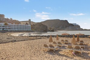 A beach with lounge chairs and umbrellas, a rocky coastline, and a hotel in the background.