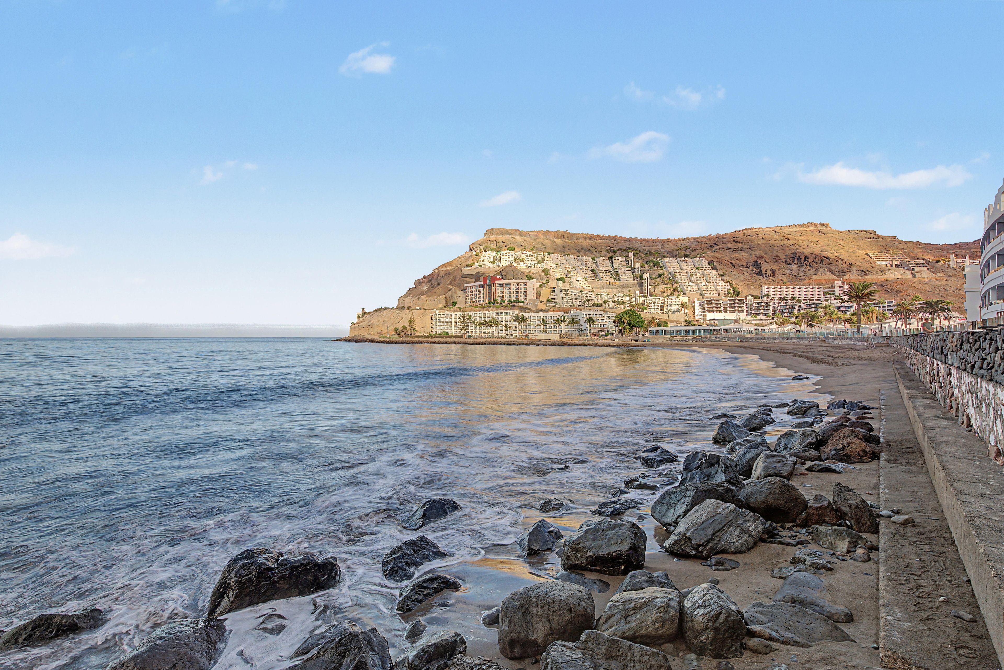Eine Küstenstadt mit Häusern an einem Hügel, einem Strand mit Felsen und ruhigem Meer.