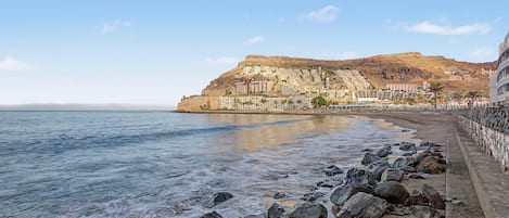 Eine Küstenstadt mit Häusern an einem Hügel, einem Strand mit Felsen und ruhigem Meer.