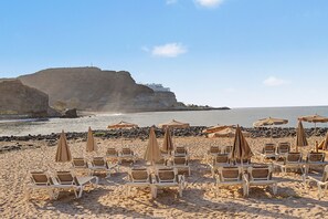 A beach with rows of sun loungers and umbrellas, a rocky cliff in the background, and calm sea. - Views of Playa del Cura with Fantastic Terrace (Mogán)