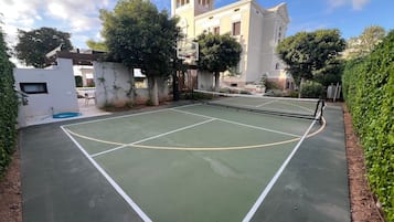 An outdoor tennis court with a net, surrounded by greenery and a building with a red roof.