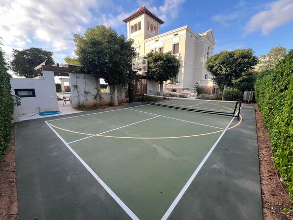An outdoor tennis court with a net, surrounded by greenery and a building with a red roof.