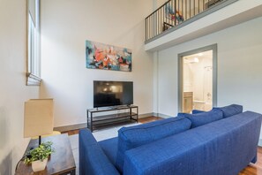 A living room with a large blue sofa, a flat-screen TV on a stand, a painting on the wall, and a staircase leading to an upper level. - Designer Loft - Parking Nearby Hospitals - 2F (Louisville)