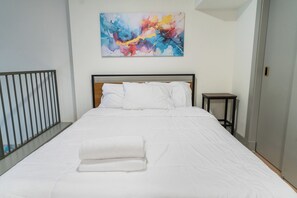 A neatly made bed with white linens and a folded white towel, a small bedside table, and a colorful abstract painting on the wall. - Designer Loft - Parking Nearby Hospitals - 2F (Louisville)