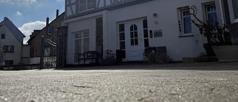 Een traditioneel vakwerkhuis met een hekpoort, een klein terras en een heldere lucht met zonlicht.