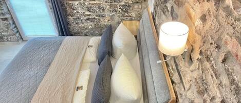A bed with a headboard against a stone wall, a bedside lamp, and a window with blinds.