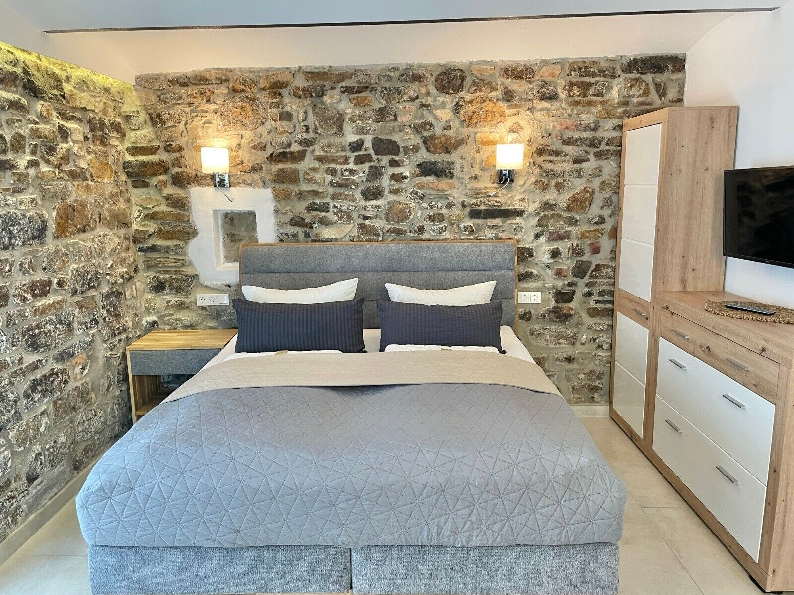 A bedroom with a stone wall, a bed with a grey quilt, two bedside lamps, and a TV on a wooden cabinet.