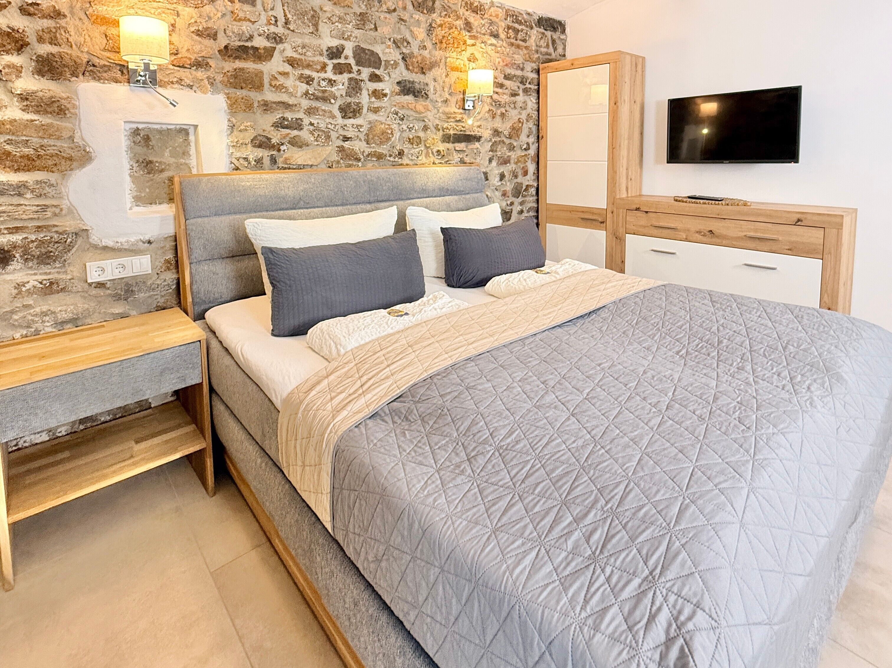 Ein Schlafzimmer mit Steinwand, einem Bett mit gesteppter Bettdecke, Nachttischen, einem Fernseher und einem Holzschrank.
