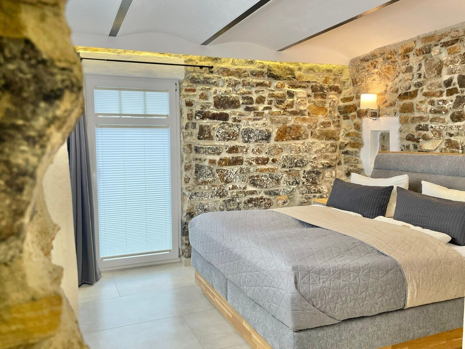 Una camera da letto con parete in pietra, un letto con copripiumino grigio e una finestra con persiane.