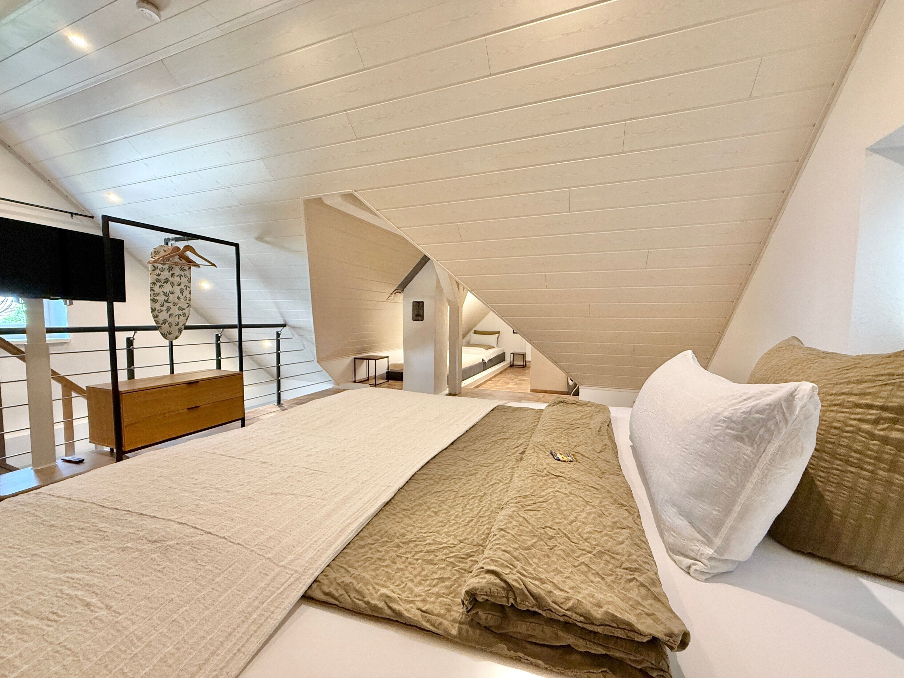 Une chambre avec un plafond en pente, un lit recouvert d’une courtepointe beige, un oreiller blanc et un banc en bois.
