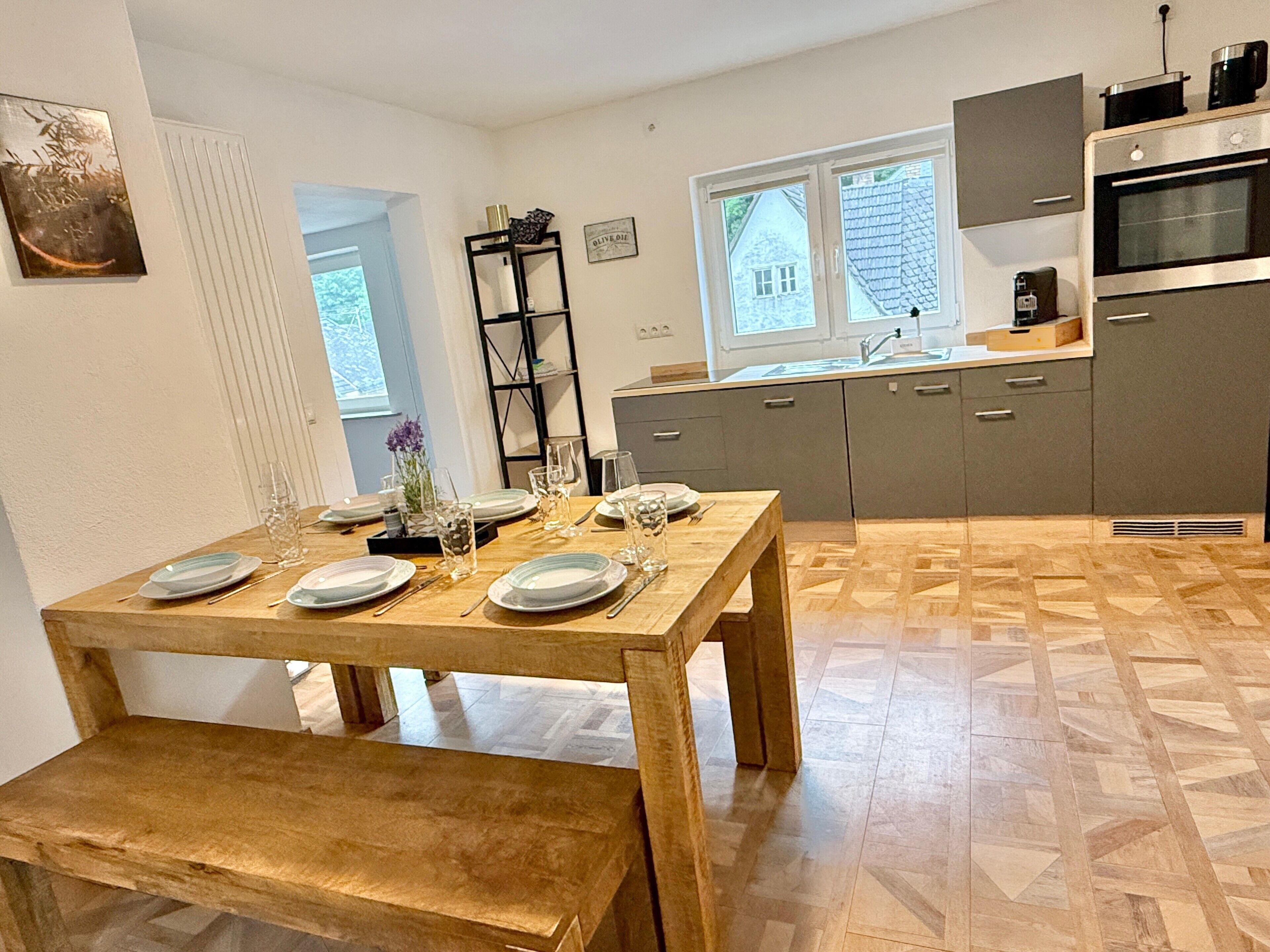 A dining area with a wooden table set for four, a bench, and a kitchen in the background with a window and a shelf.
