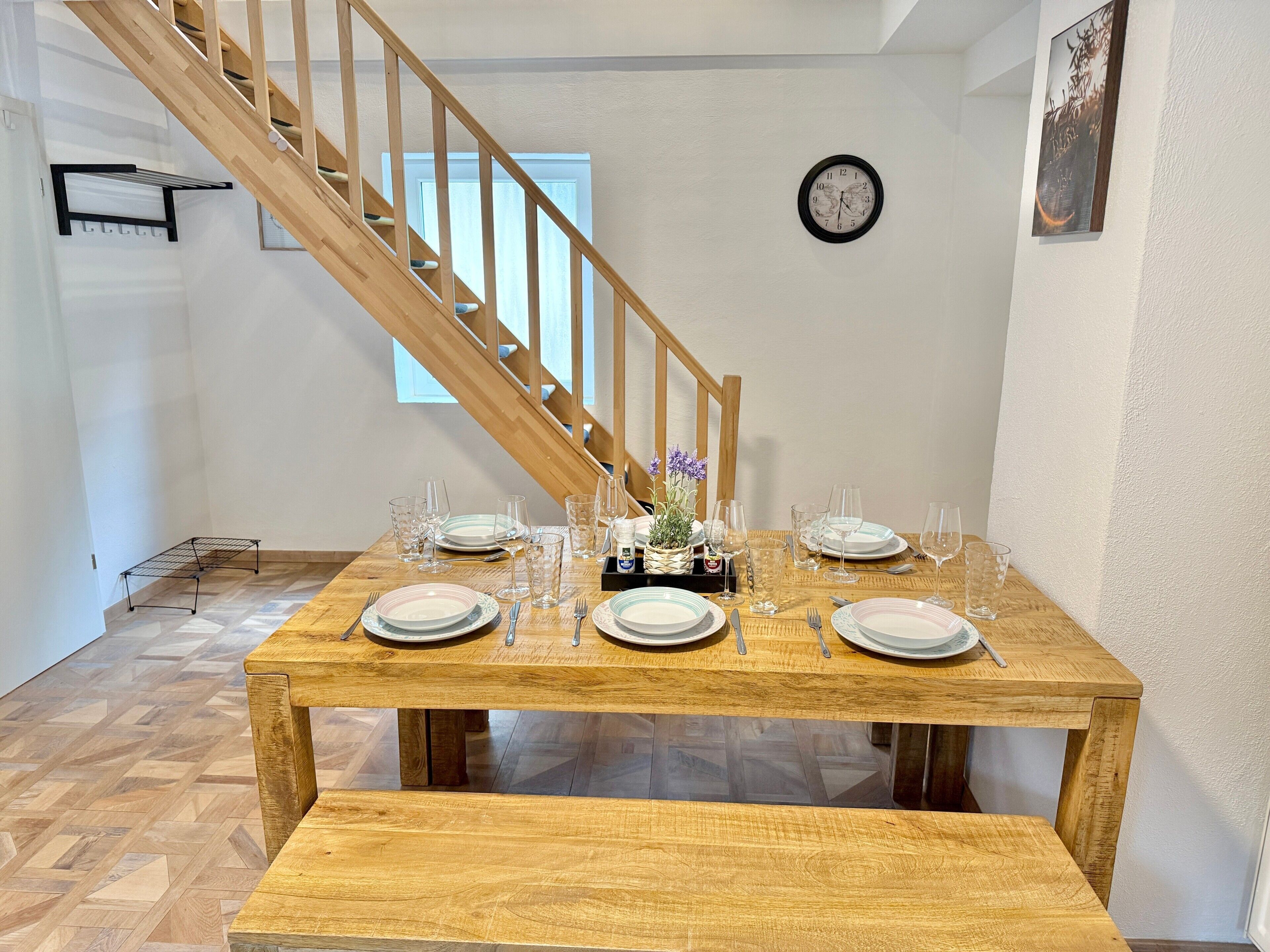A dining area with a wooden table set for four, a bench, a staircase, a clock, and a window.