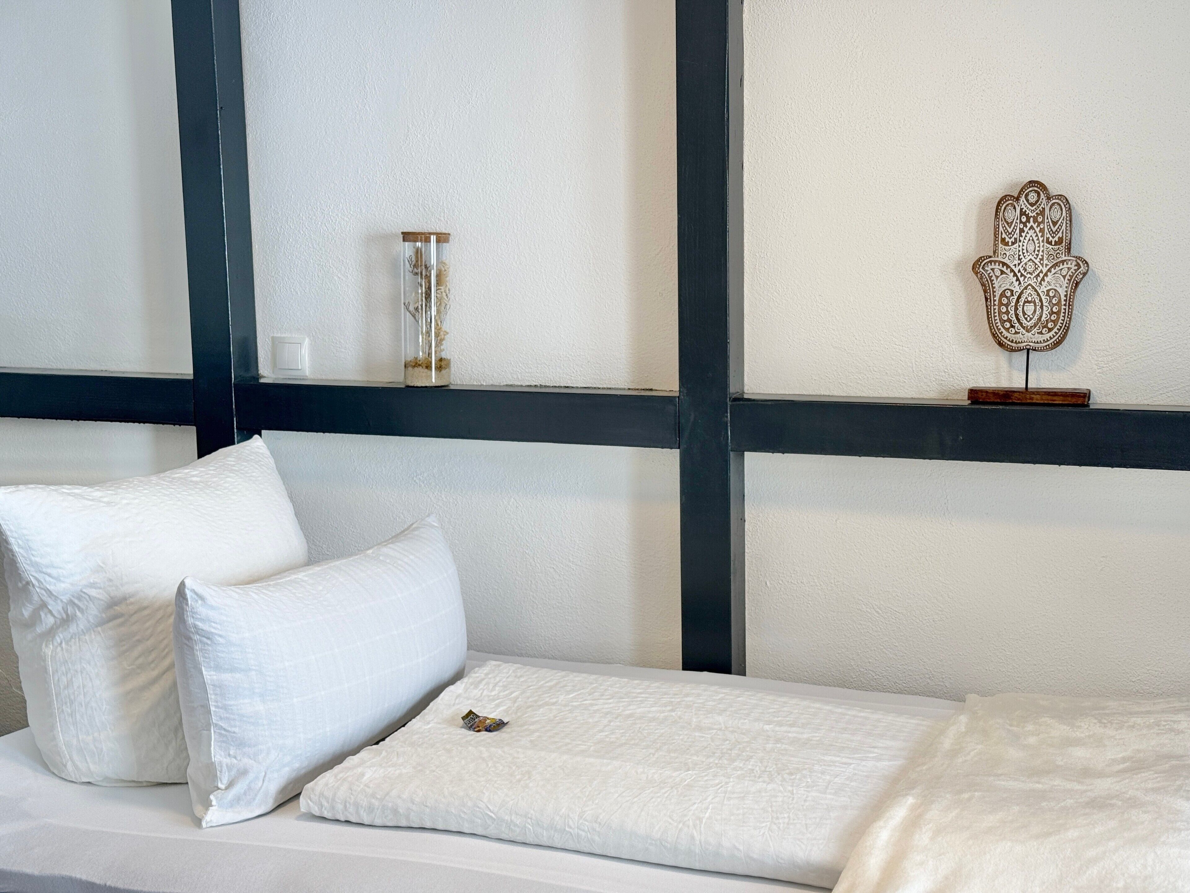A bed with white bedding and pillows, a small decorative item on the bed, and a wall with a black beam and a decorative wall piece.