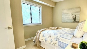 A bedroom with a bed, a window, a painting, a bedside table, and a door. - Serene Villa Steps From Beach w/ Pool and Tennis! (Hilton Head Island)