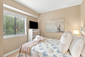 Ein Schlafzimmer mit einem Bett, einem Fernseher, einem Fenster mit Jalousien, einem Bild und einer Nachttischlampe.