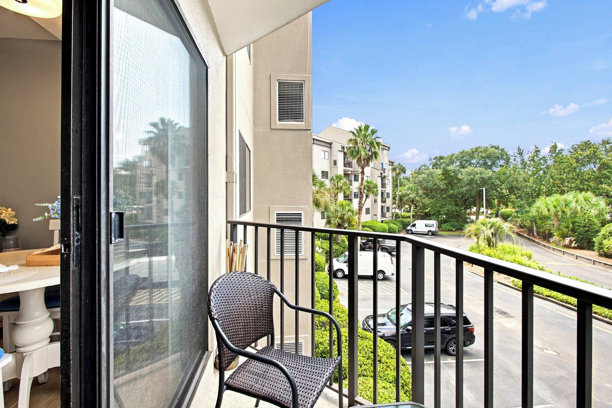 Woven wicker seating sits ready for your morning coffee on this breezy, sun-drenched balcony. The glass door opens wide to connect the cozy interior with leafy views and a bright, expansive sky.