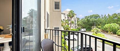 Woven wicker seating sits ready for your morning coffee on this breezy, sun-drenched balcony. The glass door opens wide to connect the cozy interior with leafy views and a bright, expansive sky.