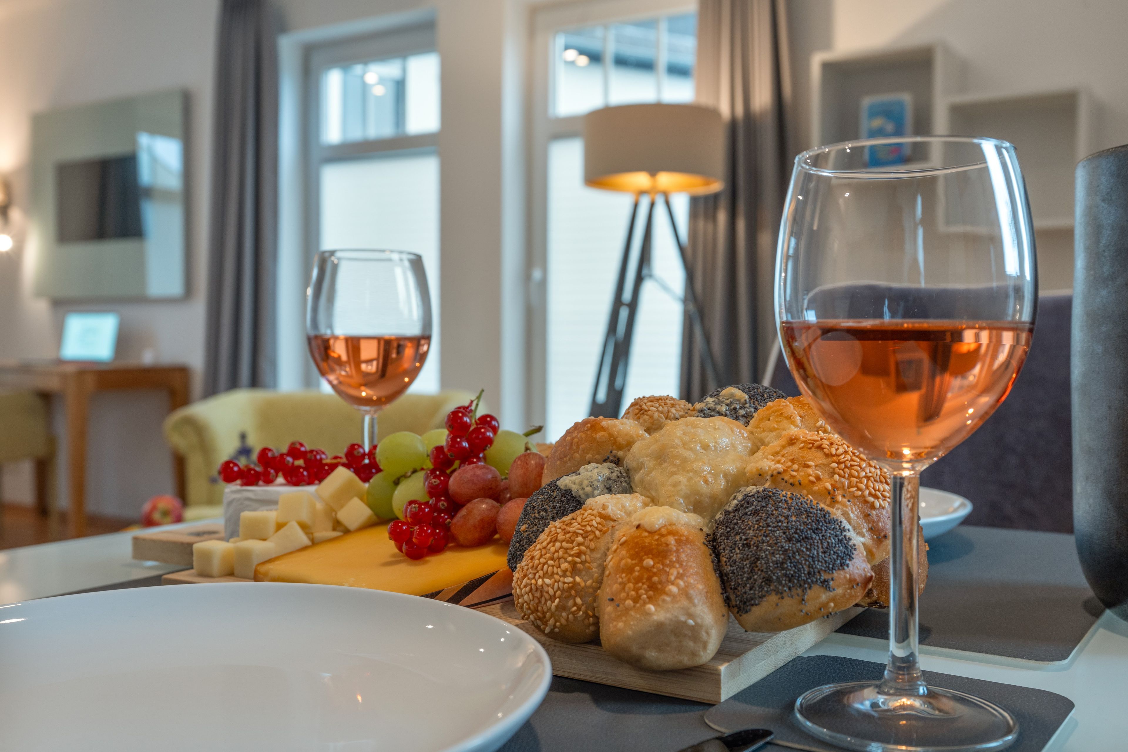 Et spisebord dækket med et udvalg af oste, brød og frugter, samt to glas rosévin.