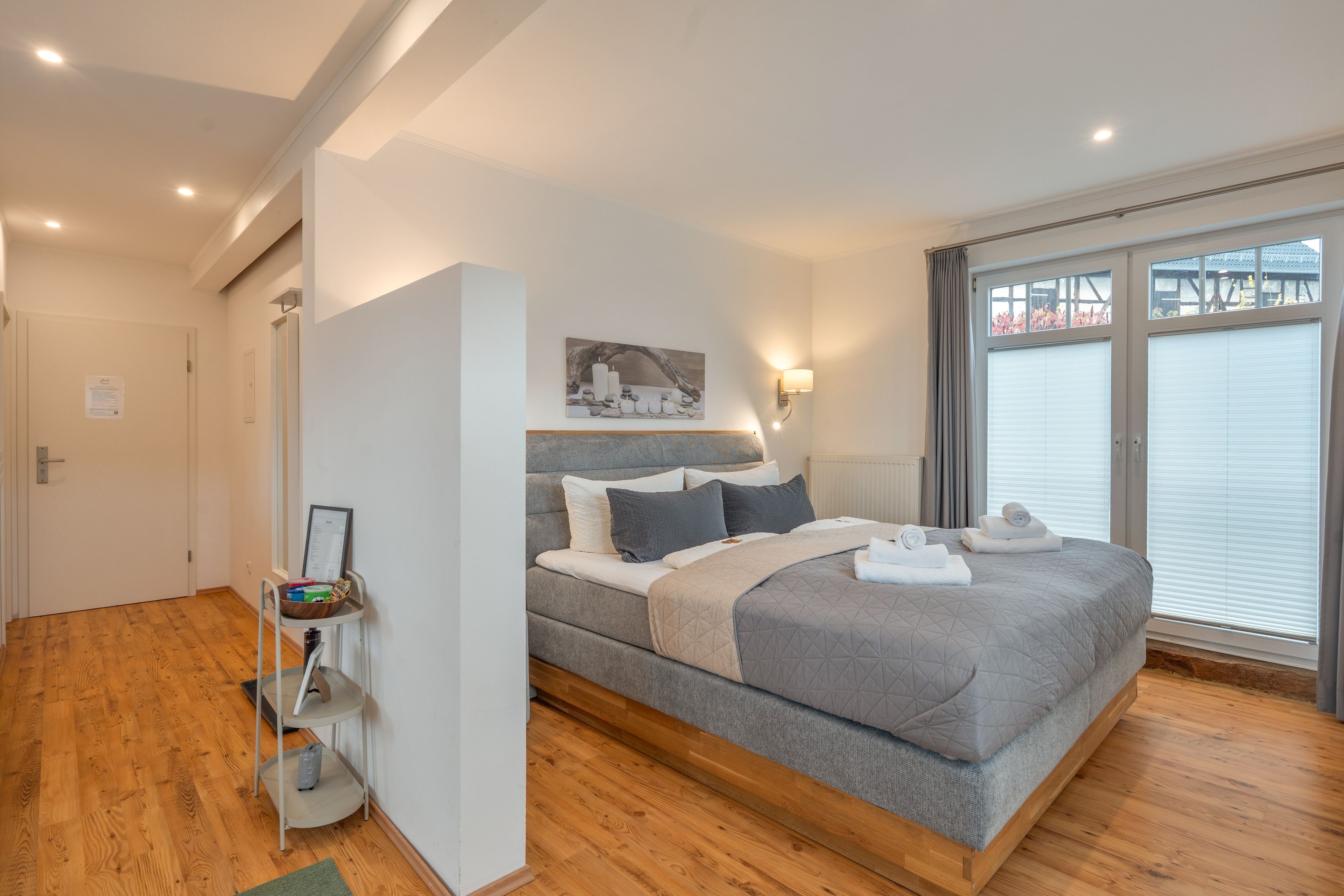 A modern bedroom with a large bed, a bedside table, a wall-mounted light, and a window with blinds.
