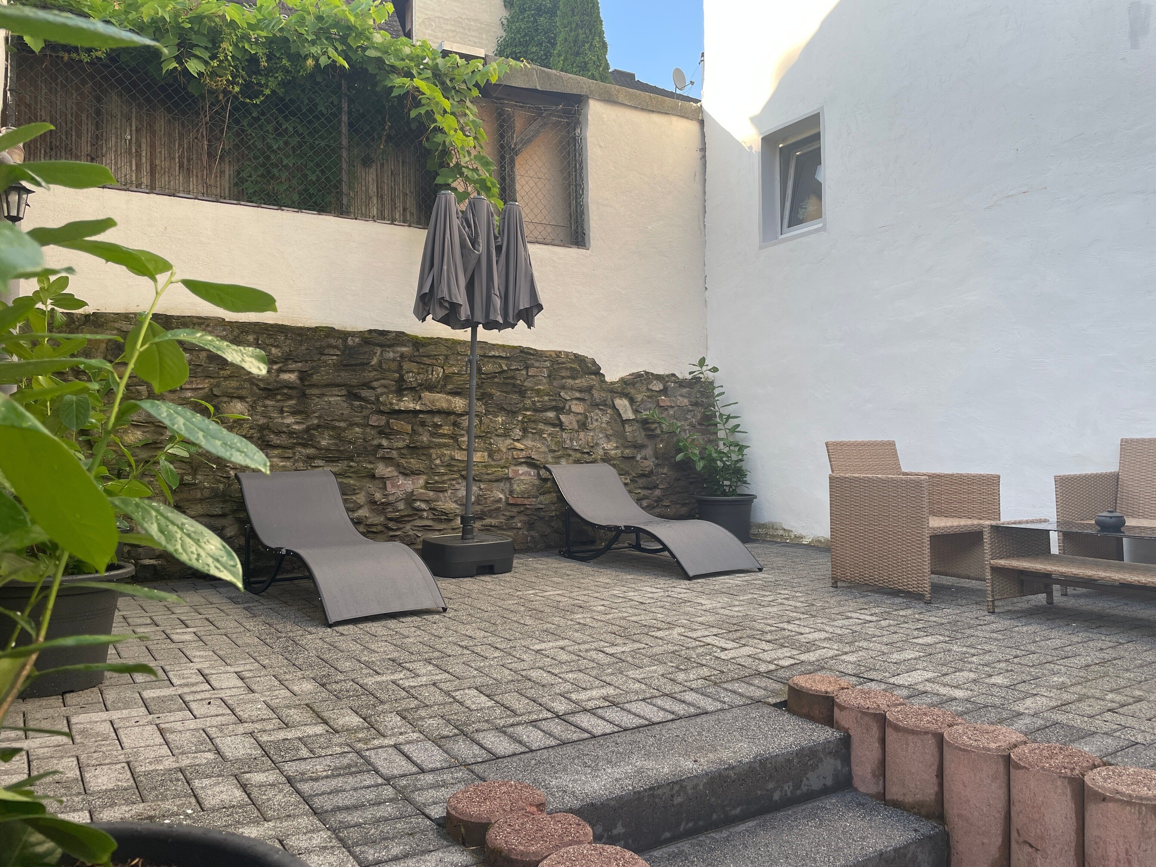 Een terras buiten, met een stenen muur, een zithoek met lounge fauteuils en een potplant.