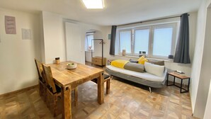 Ein gemütlicher Wohnraum mit Esstisch aus Holz, einer Couch mit Kissen, einem Fenster mit Jalousien und einer Deckenleuchte.