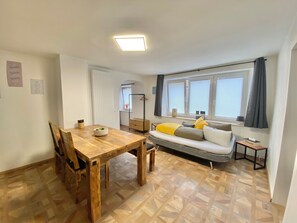 A compact living space with a wooden dining table, a sofa with cushions, a window with blinds, and a ceiling light fixture. - Apartment 'Ferienwohnung Kleinôd' with Private Terrace and Wi-Fi (Bendorf)