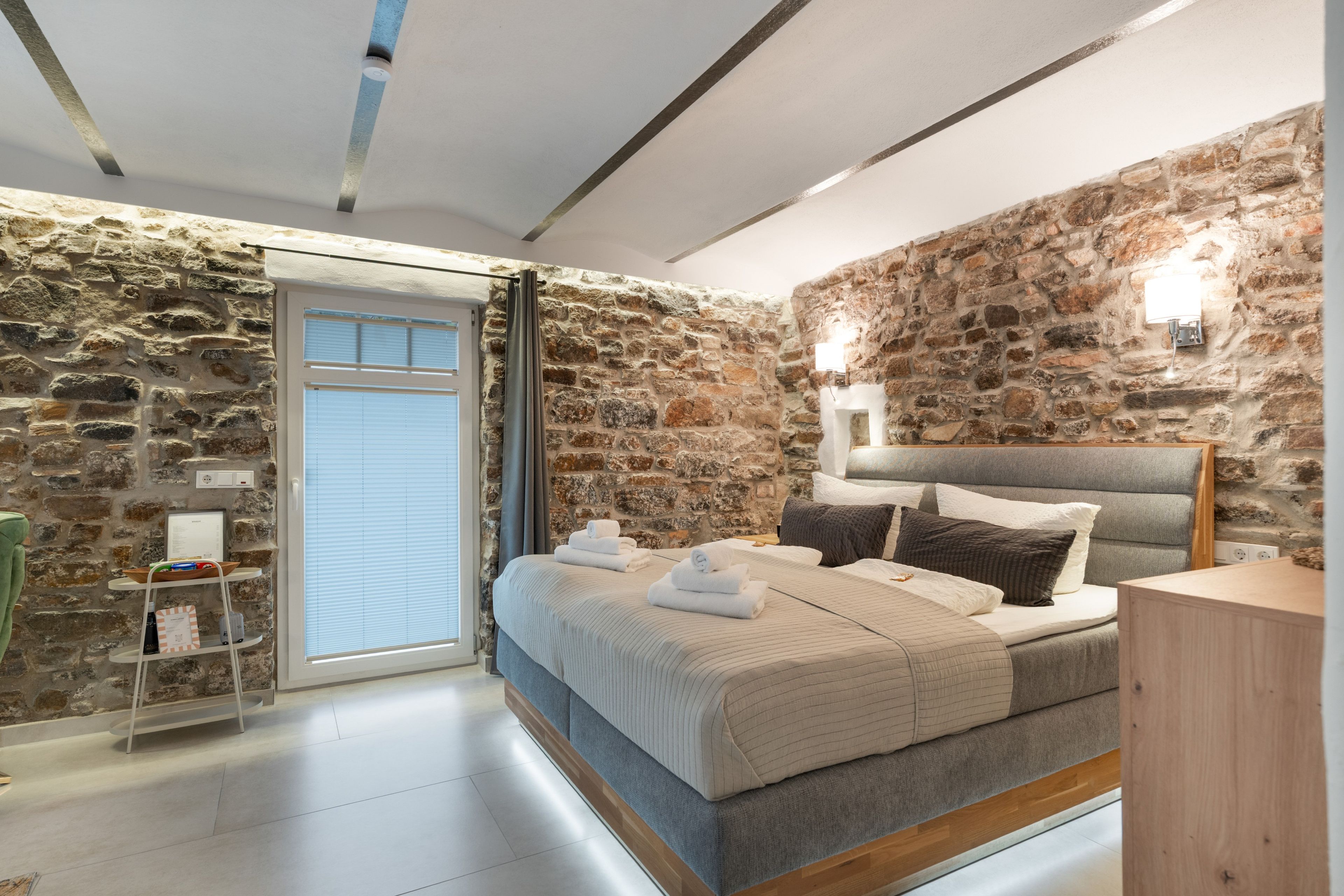A bedroom with a stone wall, a bed with towels, a bedside table, and a nightstand.