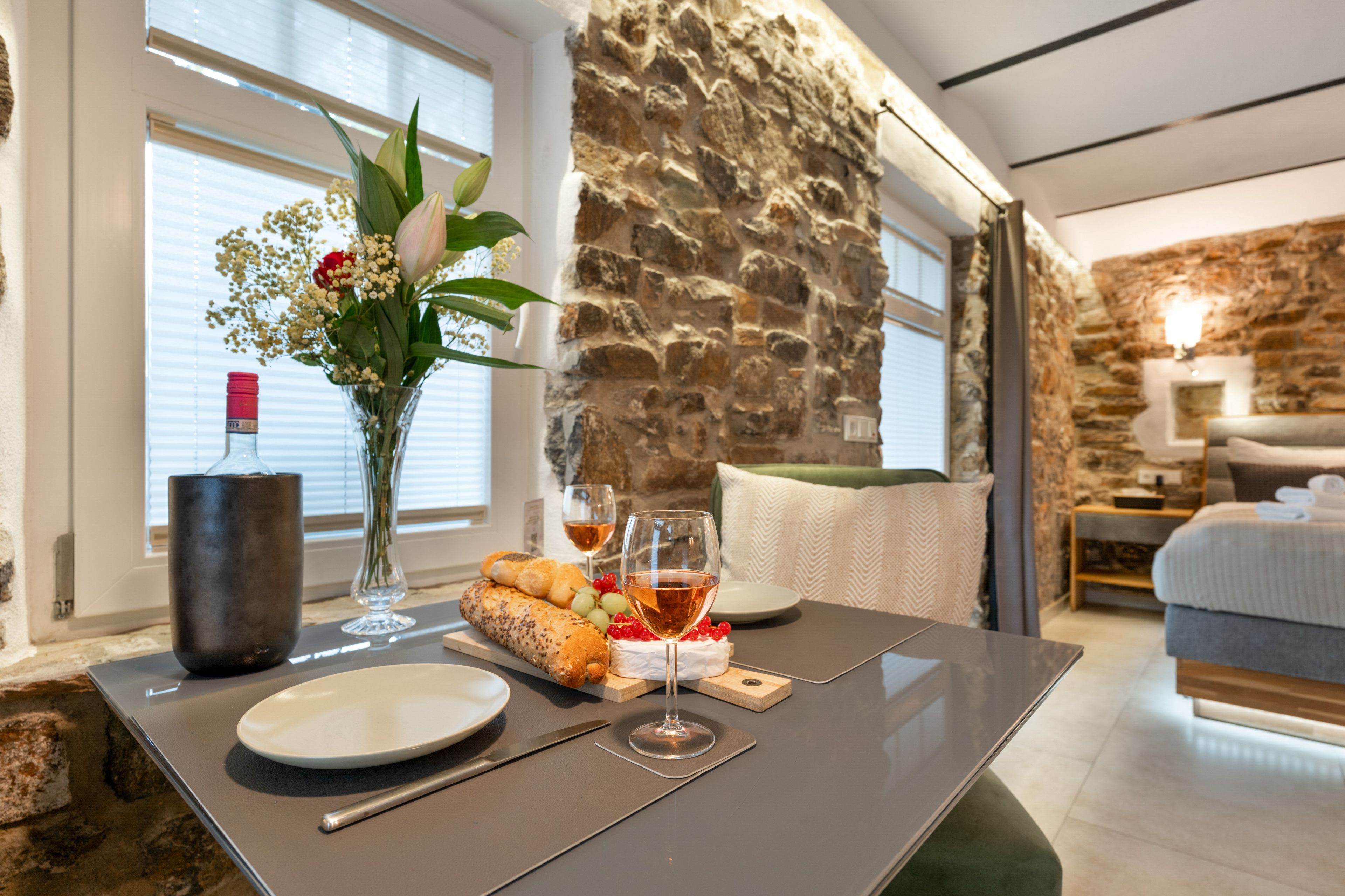 A dining area with a table set for two, featuring a bottle, glasses, bread, and a vase with flowers.