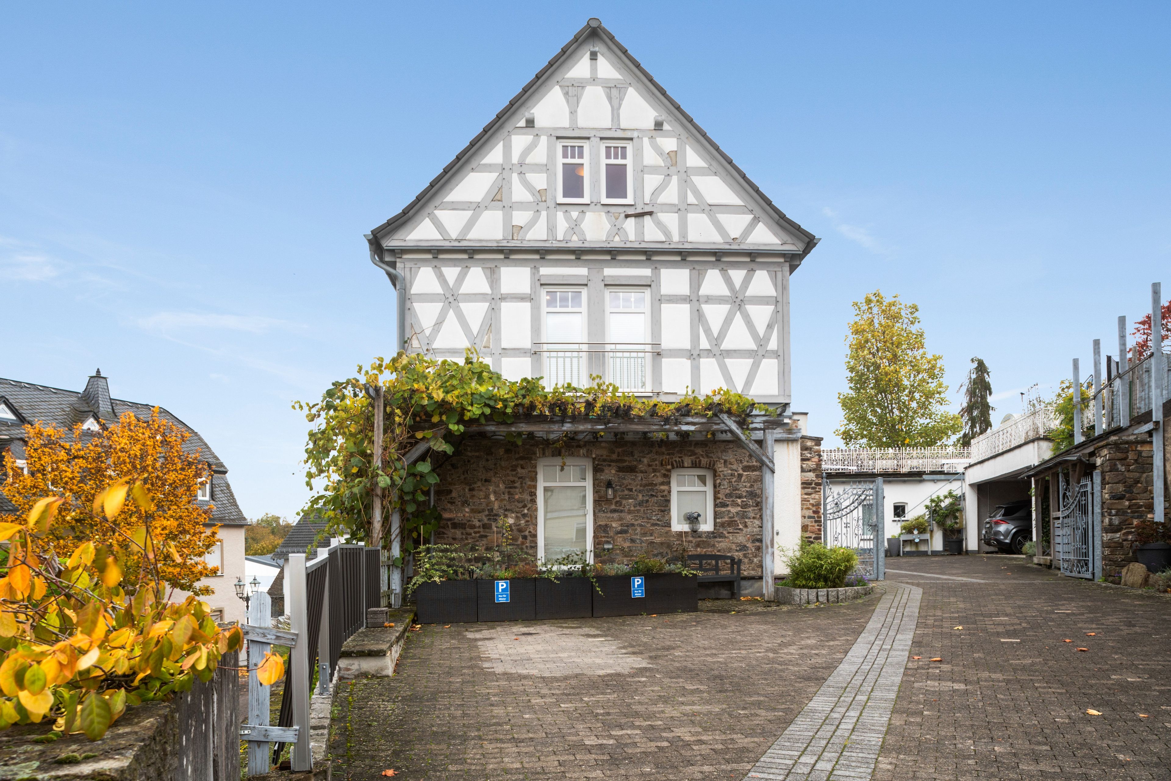 A traditional half-timbered house with a stone extension, a paved driveway, and a parking area with blue signs.