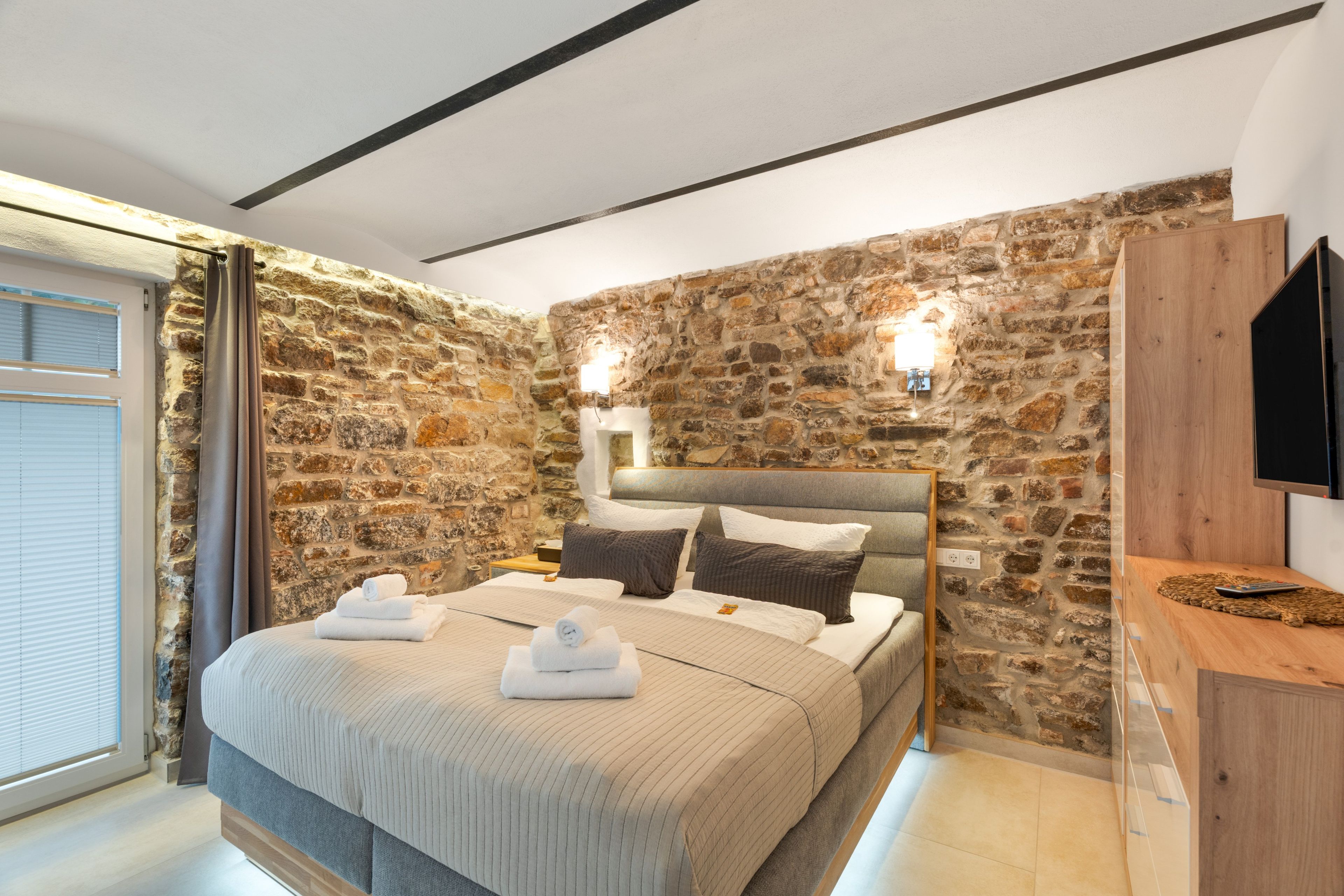 A bedroom with a stone wall, a bed with linens, a bedside table, a TV, and a window with blinds.